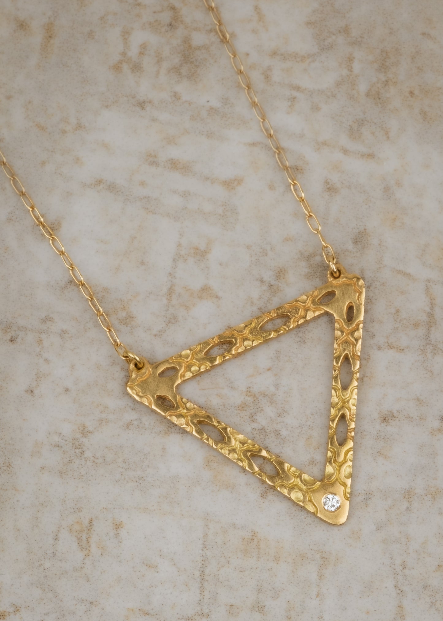 Like two souls finding their reflections in one another, the Tryst necklace creates the illusion of mirrored triangles in a celebration of negative space. Dangling from a delicate chain, hand-carved textured gold is dotted with open oblongs that draw the eye to the nadir of the triangle, where a sparkling diamond punctuates with brilliance.