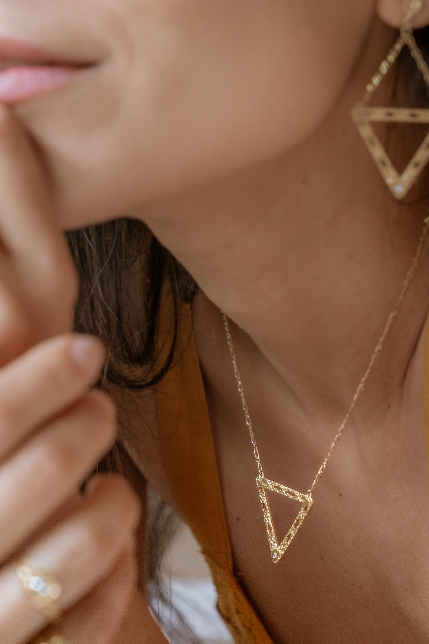 Like two souls finding their reflections in one another, the Tryst necklace creates the illusion of mirrored triangles in a celebration of negative space. Dangling from a delicate chain, hand-carved textured gold is dotted with open oblongs that draw the eye to the nadir of the triangle, where a sparkling diamond punctuates with brilliance.