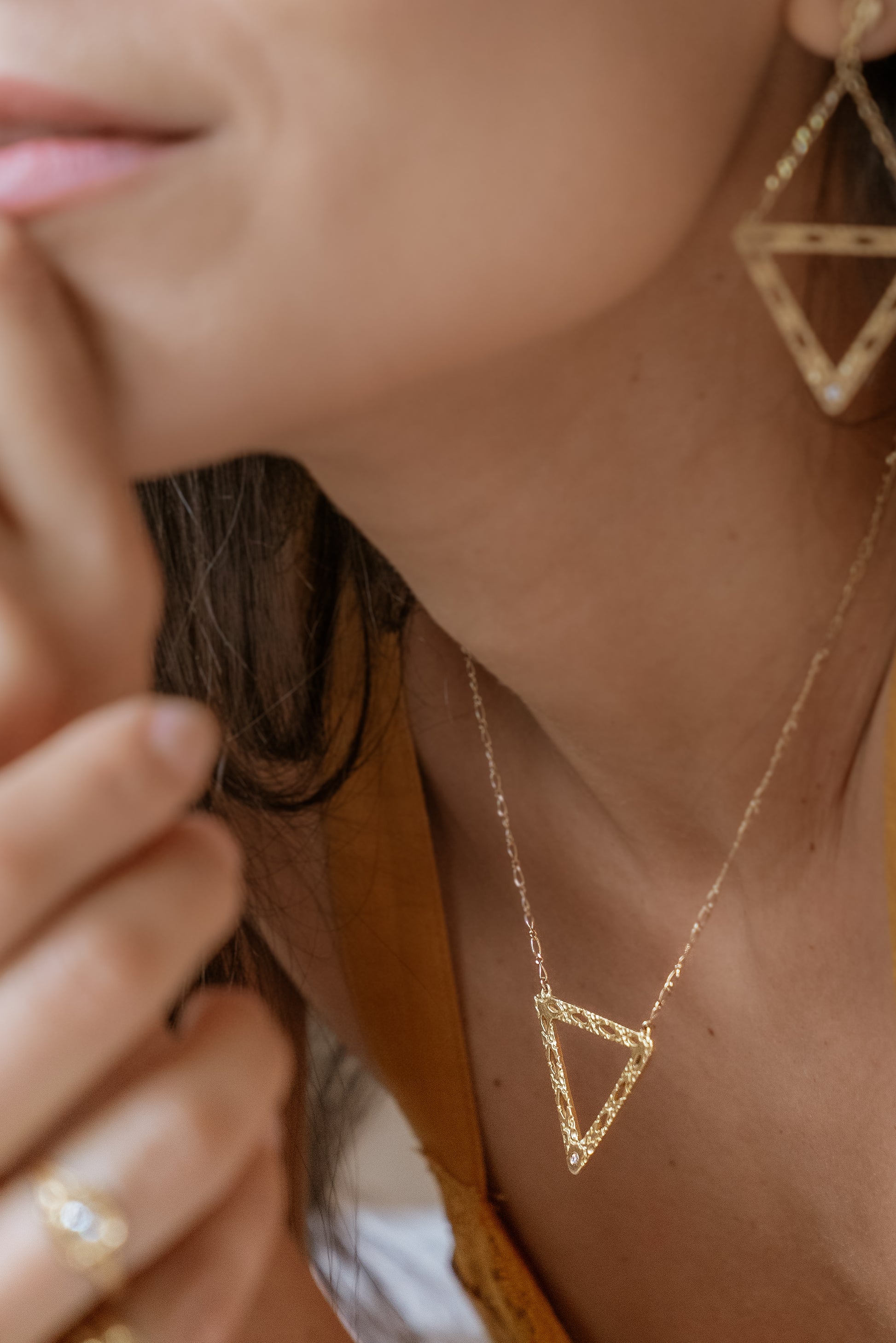 Like two souls finding their reflections in one another, the Tryst necklace creates the illusion of mirrored triangles in a celebration of negative space. Dangling from a delicate chain, hand-carved textured gold is dotted with open oblongs that draw the eye to the nadir of the triangle, where a sparkling diamond punctuates with brilliance. 